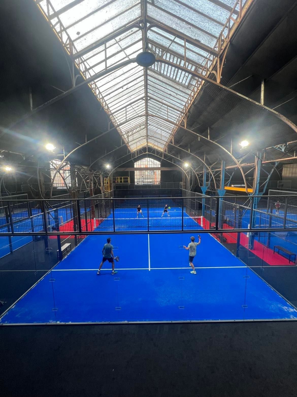Four people playing padel tennis inside an industrial-style indoor court with blue flooring.